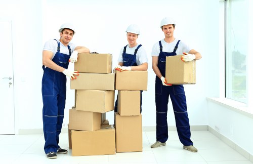 Three professional movers wearing white safety helmets, blue overalls, and white gloves are standing inside a bright, modern residential property with white walls and large windows, which allows natural light to fill the space. They are positioned around a stack of seven cardboard boxes, some stacked vertically and others placed side by side, all sealed with packing tape. The boxes vary in size, with some larger and rectangular, and others smaller. The mover on the left is holding a box with both hands, while the one in the middle, also holding a box, is standing behind the stacked boxes, and the third mover on the right is lifting a medium-sized box, preparing for loading or unloading. The flooring appears to be light-colored laminate or tile, contributing to the clean, professional atmosphere. The window on the right showcases a glimpse of a residential street in Soho, indicating a city centre location. All individuals are focused and appear to be in the process of a house relocation, with the scene illustrating the careful handling and transport of household items by Soho Man with Van’s removals service, suitable for clients in the Soho or nearby London area.