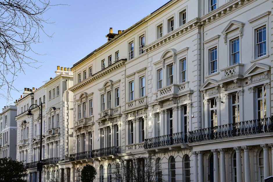 A row of multi-storey residential buildings on a narrow cobblestone street, with each property featuring distinct facades painted in neutral and pastel colors. The buildings have multiple small balcon